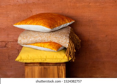 Stock of colorful pillows on wood chair against textured terracotta wall. Pile of soft yellow and orange cushions and blanket on stool near wall with space for text. Modern and cozy home concept.