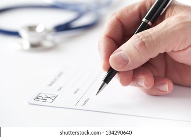Stock image with a doctor's hand writing a prescription on a Rx blank form. Nearby is seen his stethoscope on the table.