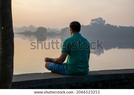Stock photo of 30 to 40 year old Indian man practicing pranayama or yoga or meditation by the lake sitting in lotus pose at Rankala lake, Kolhapur, Maharashtra, India. peaceful and scenic view.