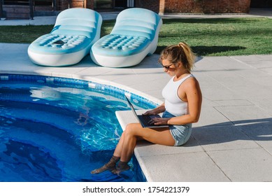 Stock photo of a caucasian woman sitting on the poolside with her feet in the water. She is typing on her laptop. She is wearing sunglasses.