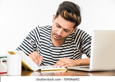 Stock Photo Of Handsome Indian Boy / Male College Student Studying On Study Table With Pile Of Books, Laptop Computer And Coffee Mug. Smiling Or Thinking/worried/showing Thumbs Up / With Smartphone
