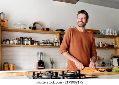 Stock photo of a middle aged man holding a knife and slicing up some vegetables in a kitchen. He is looking through a window and smiling. - Powered by Shutterstock