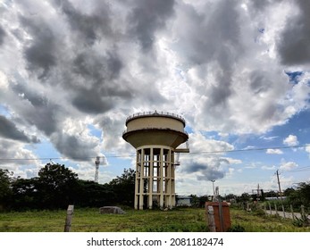 Stock photo old rusty yellow painted water tank tower surrounded by green trees under blue sky and white clouds at the middle the land  small power house shelter near the water tank at Gulbarga