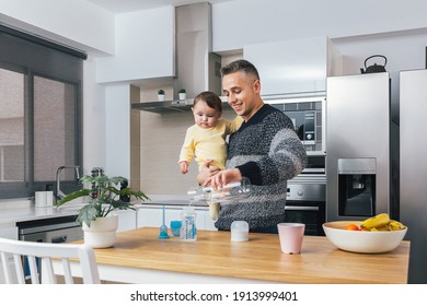 Stock Photo Of Young Single Father Preparing A Feeding Bottle While Holding His Baby In Arms In The Kitchen. Man And Child, Parenting Fatherhood Real Moment. Single Dad Family Home Life Concept