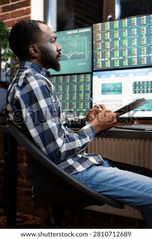Stockbroker writes notes while studying forex market trends on multiple monitors at night, reviewing financial data and stock charts, with chart analysis and trading workspace context.