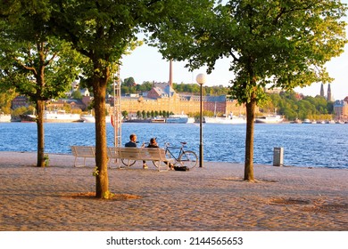 Stockholm, Sweden - June 26, 2019: Romantic Couple Sitting On A Bench Among The Green Trees In Front Of Blue Water Of Riddarfjärden On Riddarholmen Island With A View To Södermalm