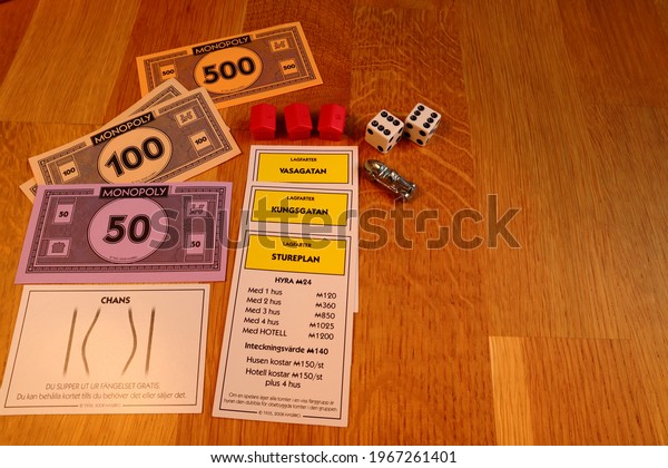 Stockholm, Sweden. May 02, 2021. Three yellow\
monopoly street card against wooden background. Classical board\
game. Swedish text for Vasagatan, Stureplan, Kungsgatan. Red hotel,\
money, dice and car.