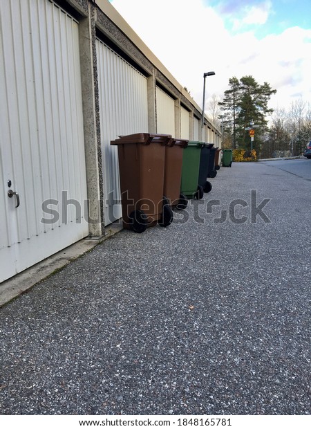 Stockholm, Sweden. November 02, 2020. A row of brown and\
green trashcans outside. Next to a gray wall of carports. Litter,\
dumping in the bins. Asphalt on the ground and some clouds in the\
sky. 