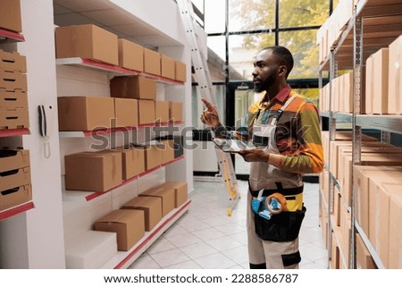 Stockroom manager in industrial overall checking boxes, working at customers order preparing products for delivery in storehouse. African american worker managing parcels transportation in warehouse