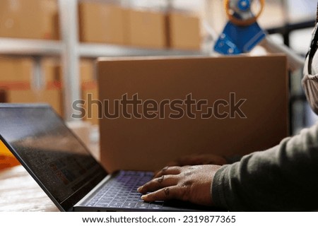 Stockroom worker checking goods quality control report on laptop computer before start working at customers order in warehouse. African american employee preparing packages for delivey. Close up