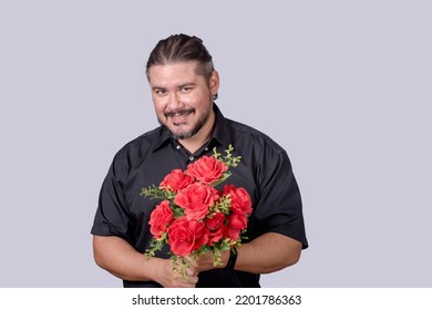 A stocky bearded man in a black polo shirt holding a bouquet of red roses. A single male in his 30s wooing and impressing a date. Isolated on a gray background.