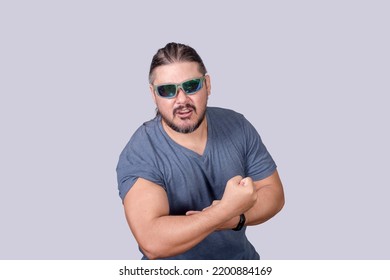 A stocky and burly man wearing shades raises his sleeve to expose his arms, flexing his biceps. Grunting while trying to look big.