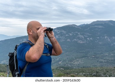 A stocky Caucasian hiher observing the landscape with binoculars on a gray and cloudy day. 