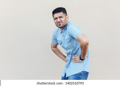 Stomach Pain Or Diet Problem. Portrait Of Sick Handsome Young Bearded Man In Blue Shirt Standing And Holding His Painful Belly, Feeling Bad. Indoor Studio Shot Isolated On Light Beige Wall Background.