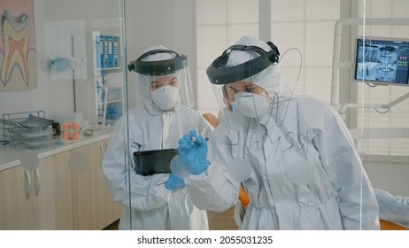 Stomatological Doctor Drawing Tooth On Glass With Marker Explaining Dental Care Procedure And Operation To Patient Sitting In Cabinet. Dentist And Nurse With Virus Protection Suits