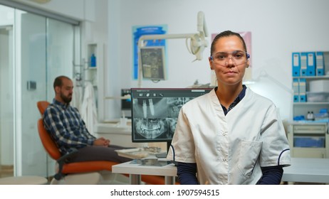 Stomatologist Doctor Talking On Web Cam With Patients About Oral Hygiene Looking At Camera While Patient Waiting For Nurse In Background. Dental Assistant Preparing Man For Stomatological Surgery.