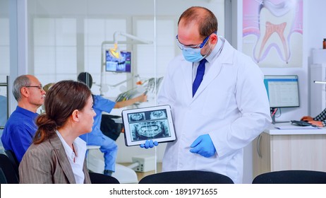 Stomatologist Pointing On Digital Screen Explaining X-ray To Woman Sitting On Chair In Waiting Room Of Stomatological Clinic. Doctor Working In Modern Dental Clinic, Examining, Showing Radiography