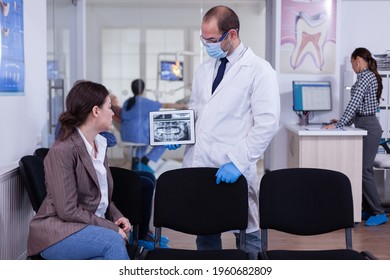 Stomatologist Pointing On Digital Screen Explaining X-ray To Woman Sitting On Chair In Waiting Room Of Stomatological Clinic. Doctor Working In Modern Dental Clinic, Examining, Showing Radiography.