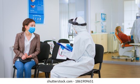 Stomatologist In Protective Suit Reviewing Registration Form With Patient Explaining Treatment Writing On Clipboard In Covid-19 Pandemic. Medical Nurse Wearing Face Shield, Coverall, Mask And Gloves.