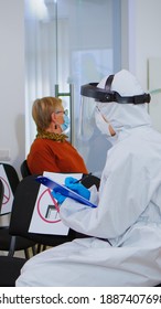 Stomatologist In Protective Suit Reviewing Registration Form With Patient Explaining Treatment Writing On Clipboard In Covid-19 Pandemic. Medical Nurse Wearing Face Shield, Coverall, Mask And Gloves.