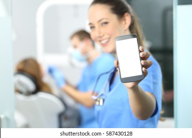 Stomatologist showing phone screen and looking at you in a dentist office interior with a doctor working in the background