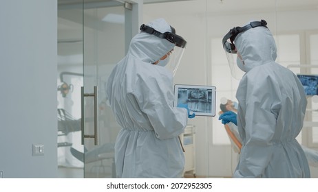 Stomatologists With Ppe Suits Holding Digital Tablet With Teeth X Ray Analyzing Dental Healthcare For Implant Operation At Medical Clinic. Dentists Using Scan Technology During Pandemic