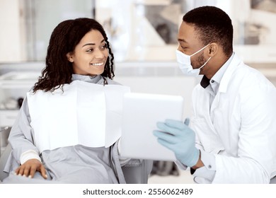 Stomatology Concept. Black male dentist in protective medical mask showing teeth picture on digital tablet to female patient during check up in dental clinic, doctor discussing treatment with woman - Powered by Shutterstock