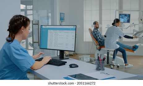 Stomatology nurse sitting at desk working on computer for appointments at dental clinic. Assistant using monitor screen while dentist doing professional examination in background