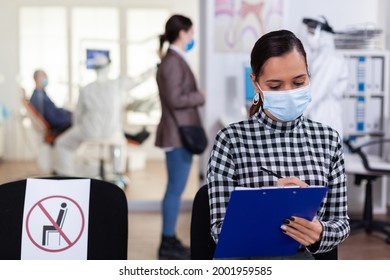 Stomatology Patient In Dentistry Waiting Area Filling Form Before Consultation With Dentist Dressed In Ppe Suit As Safety Precation Against Infection With Coronavirus During Global Outbreak.