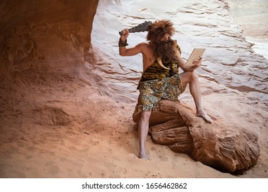 Stone Age luddite caveman scratching his head with a club while looking at his stone tablet outdoors in a weathered rock cave