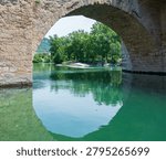 Stone arch bridge reflected in the emerald waters of the Tarn River in Millau, southern France. Historic masonry architecture creating a natural geometric frame and tranquil riverscape.