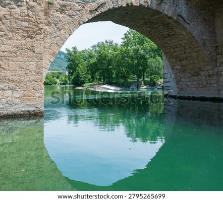 Stone arch bridge reflected in the emerald waters of the Tarn River in Millau, southern France. Historic masonry architecture creating a natural geometric frame and tranquil riverscape.