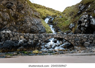 A stone arch spanning over a rapid stream, flowing downwards, captured from a sandy beach in Scotland, with wet sand visible in the lower part of the image. - Powered by Shutterstock