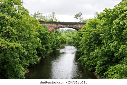 A stone bridge arches over a serene river Irwell Salford surrounded by lush greenery on a calm, cloudy day. - Powered by Shutterstock