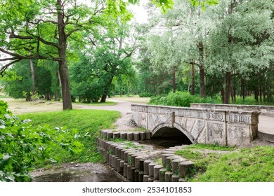 A stone bridge arches over a small stream in a serene park, surrounded by tall, green trees with a winding path leading toward the bridge. - Powered by Shutterstock