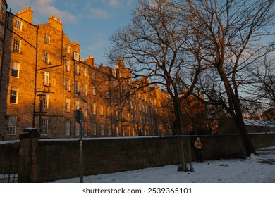 Stone buildings glow in sunset; leafless trees cast intricate shadows on snowy ground below. - Powered by Shutterstock