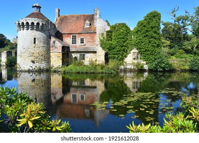 A Stone Centuries Old British Medieval Castle Facade Amongst Lush Greenery With Its Reflection In Water. It Includes A Tower Residential Buildings And An Estates Office All Dominated By A Banquet Hall