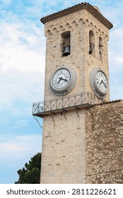 Stone clock tower with arched windows and large round clocks on its facade, set against a blue sky in Cannes, France. Part of a historic building, trees visible in background.
