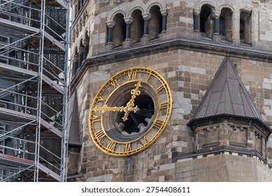 Stone clock tower features a large golden clock face with Roman numerals. Arched windows sit above the clock. Scaffolding lines the left side of the tower