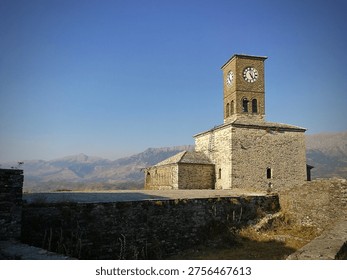 Stone clock tower situated within Gjirokaster Castle in southern Albania. The structure features a square tower with a clock face on all sides, built from traditional stonework. The surrounding landscape includes mountainous terrain under a clear blue sky, contributing to the historical ambiance. The castle itself is a historic landmark, offering a glimpse into Albania's rich history and architectural heritage. The foreground includes a stone wall and sparse vegetation.