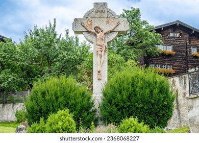 A stone crucifix of Christ stands amidst green plants near Parish church of St.Georg in Ernen, Switzerland.