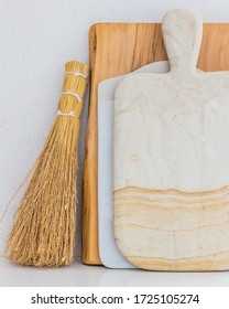 A Stone Cutting Board In Front Of A Ceramic Cutting Board Leaning Against A Raw Edge Wooden Cutting Board, All Leaning Against A Quartz Backsplash. A Straw Whisk Broom Leans Against The Side.