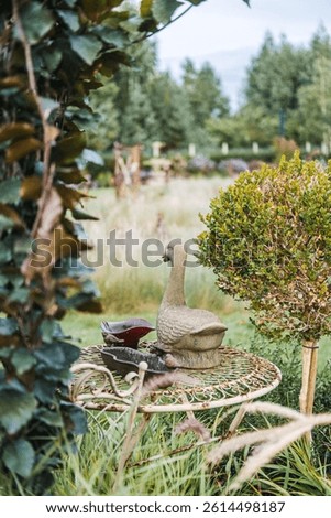 Stone duck sculpture and ceramic pieces on a vintage garden table surrounded by tall grasses and shrubs. Whimsical garden decor, rustic charm, ornamental wildlife, outdoor art. High quality photo