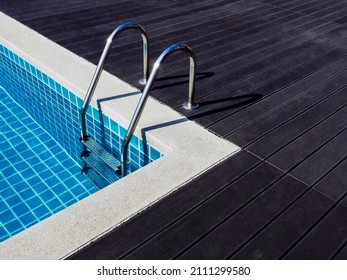 Stone Edge Frame On The Square Swimming Pool With Clean Clear Water, Nobody. Square Shaped Pool With Blue Tiles With Grab Bars Ladder On The Dark Wood Decks, No People. Summer Background.