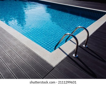 Stone Edge Frame On The Square Swimming Pool With Clean Clear Water, Nobody. Square Shaped Pool With Blue Tiles With Grab Bars Ladder On The Dark Wood Decks, No People. Summer Background.