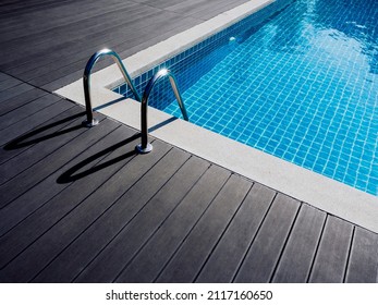 Stone Edge Frame On The Square Swimming Pool With Clean Clear Water, Nobody. Square Shaped Pool With Blue Tiles With Grab Bars Ladder On The Dark Wood Decks, No People. Summer Background.