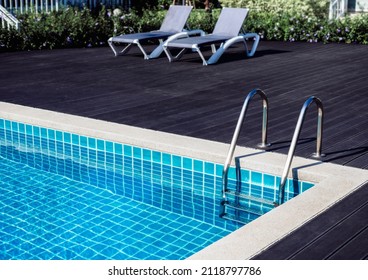 Stone Edge Frame On The Square Swimming Pool With Clean Clear Water, Nobody. Square Shaped Pool With Blue Tiles With Grab Bars Ladder And Sunbeds On The Dark Wood Decks, No People. Summer Background.