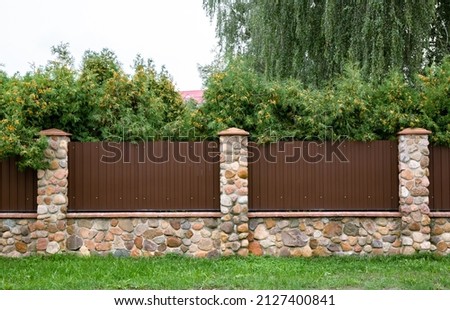 Stone fence with brown metal profile. Corrugated surface with vertical lines. Copy space. Private property fencing security. Opaque hedge. Outdoor exterior. High. Side view. Country or chalet style.