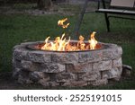 A stone fire pit with a ring of bright flames burning warmly in a backyard. The fire pit is constructed from stacked stone bricks, providing a sturdy and rustic appearance.