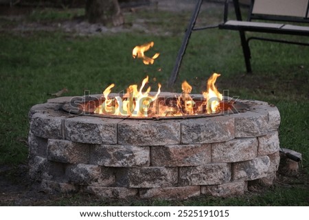 A stone fire pit with a ring of bright flames burning warmly in a backyard. The fire pit is constructed from stacked stone bricks, providing a sturdy and rustic appearance.